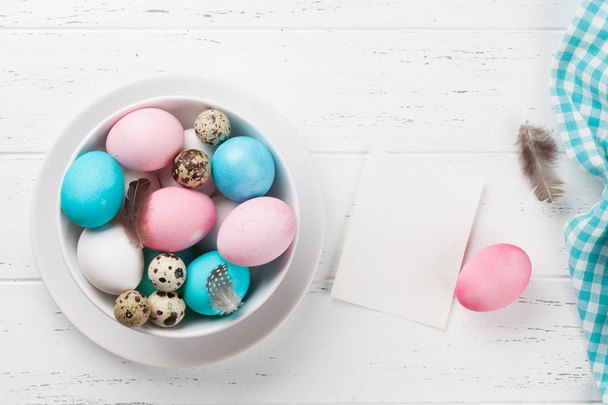 Easter greeting card with colorful eggs in bowl on white wooden table. Top view with space for your greetings - Photo, image