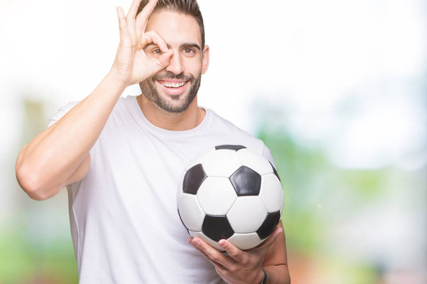 Nuori mies tilalla jalkapallo jalkapallo pallo yli eristetty tausta onnellinen kasvot hymyillen tekee ok merkki käsin silmään katselee sormien läpi
 - Valokuva, kuva