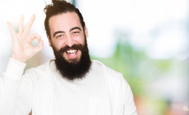 Young man with long hair and beard wearing sporty sweatshirt smiling positive doing ok sign with hand and fingers. Successful expression. - Photo, Image