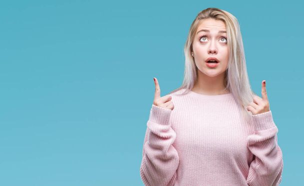 Young blonde woman wearing winter sweater over isolated background amazed and surprised looking up and pointing with fingers and raised arms. - Zdjęcie, obraz