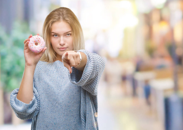 Καυκάσιος γυναίκα τρώει γλυκά ντόνατ πέρα από το απομονωμένο υπόβαθρο, δείχνοντας με το δάχτυλο στη φωτογραφική μηχανή και σε εσάς, το χέρι συνδεθείτε, θετική και σίγουρη κίνηση από το μέτωπο - Φωτογραφία, εικόνα