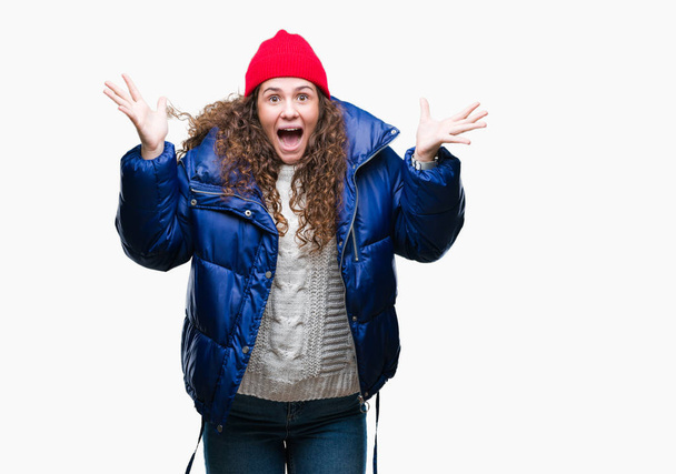 Beautiful young brunette curly hair girl wearing winter coat, wool cap and sweater over isolated background celebrating crazy and amazed for success with arms raised and open eyes screaming excited. Winner concept - Foto, Imagen