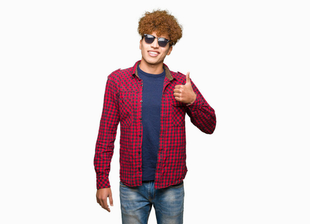 Jonge knappe man met afro haar dragen van een zonnebril gelukkig duimschroef opwaarts gebaar met de hand te doen. Goedkeuring van expressie kijken naar de camera met tonen van succes. - Foto, afbeelding