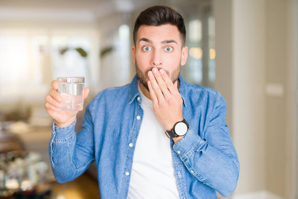 Jovem homem bonito bebendo um copo de água em casa boca tampa com a mão chocado com vergonha por erro, expressão de medo, assustado em silêncio, conceito secreto
 - Foto, Imagem