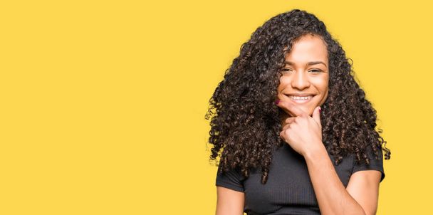 Young beautiful woman with curly hair looking confident at the camera with smile with crossed arms and hand raised on chin. Thinking positive. - Foto, Imagem