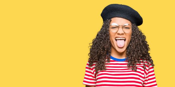 Young beautiful woman with curly hair wearing glasses and fashion beret sticking tongue out happy with funny expression. Emotion concept. - Φωτογραφία, εικόνα