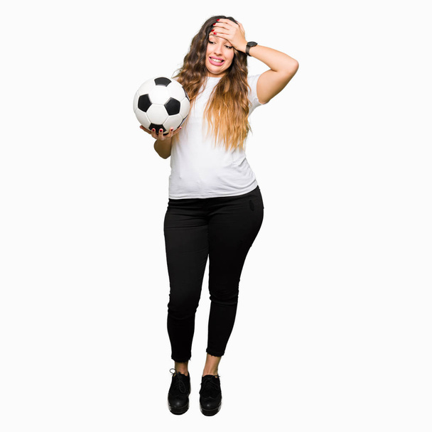 Young adult woman holding soccer football ball stressed with hand on head, shocked with shame and surprise face, angry and frustrated. Fear and upset for mistake. - Φωτογραφία, εικόνα