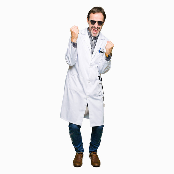 Красивый врач средних лет в солнечных очках очень счастлив и взволнован, делая жест победителя с поднятыми руками, улыбаясь и крича об успехе. Концепция празднования
. - Фото, изображение
