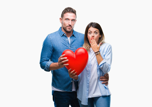 Young couple in love holding red heart over isolated background cover mouth with hand shocked with shame for mistake, expression of fear, scared in silence, secret concept - Photo, Image