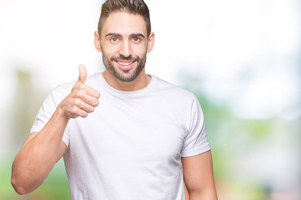 Young man wearing casual white t-shirt over isolated background doing happy thumbs up gesture with hand. Approving expression looking at the camera showing success. - Фото, изображение
