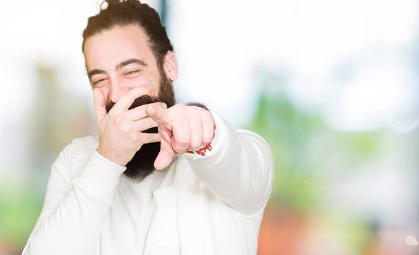 Jonge hipster man met lange haren en baard dragen sportieve sweater lachend van u, wijzend naar de camera met vinger hand over mond, schande expressie - Foto, afbeelding