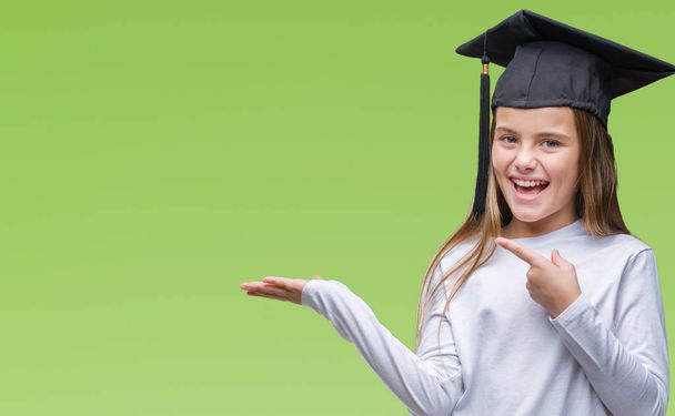 Молода красива дівчина в шапці випускника над ізольованим тлом здивована і посміхається камері, представляючи рукою і вказуючи пальцем
. - Фото, зображення
