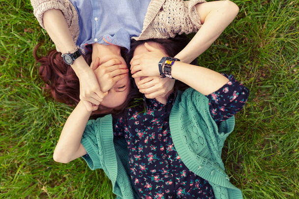 Vista superior de amigos sonrientes tumbados en la hierba divirtiéndose, cerrando los ojos con las manos. Hermanas pasando tiempo juntas
. - Foto, Imagen