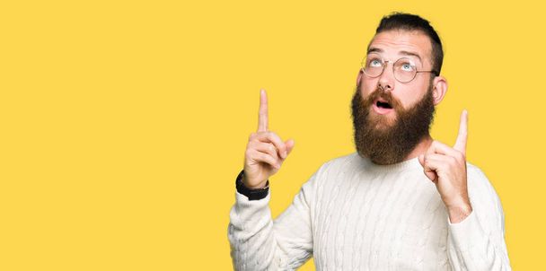 Joven hombre hipster con gafas y suéter de invierno asombrado y sorprendido mirando hacia arriba y señalando con los dedos y los brazos levantados
. - Foto, imagen
