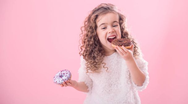 ピンクの背景、テキストのための場所に、ドーナツを食べる少しうれしそうなかわいい巻き毛の女の子の肖像画 - 写真・画像