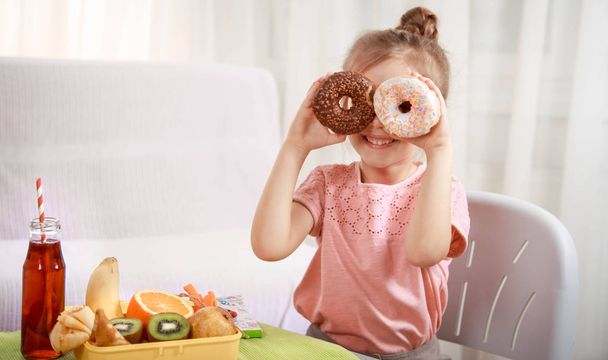 Little beautiful cheerful girl eating a donut and lunch box with fruits at home at the table, the concept of children's food - Foto, Bild