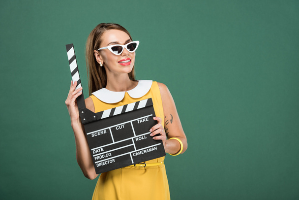 beautiful smiling stylish woman in yellow dress and sunglasses holding film clapperboard isolated on green - Photo, Image