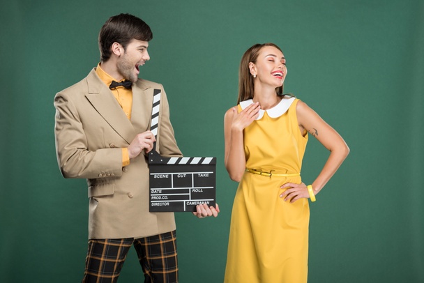 handsome man in vintage clothes holding film clapperboard while beautiful woman posing isolated on green - Φωτογραφία, εικόνα