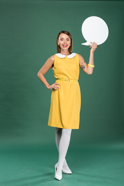 beautiful stylish woman in yellow dress looking at camera and holding speech bubble on green background - Fotografie, Obrázek