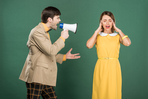 woman in vintage clothes covering ears with hands while angry man yelling in loudspeaker isolated on green - Foto, Imagen