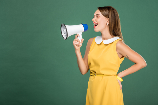 beautiful stylish woman in yellow dress shouting in loudspeaker isolated on green with copy space - Фото, изображение