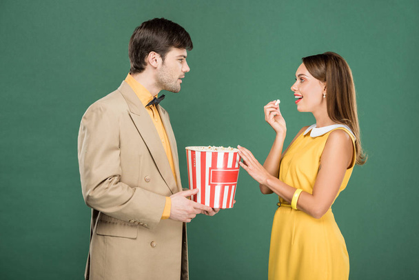 beautiful couple in vintage clothes eating popcorn and looking at each other isolated on green - Foto, Imagem
