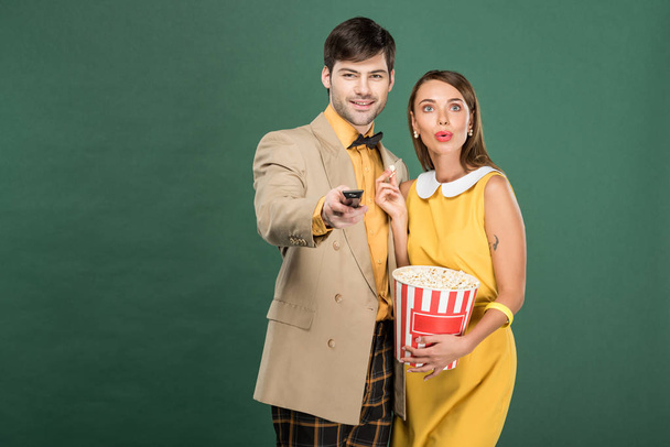 beautiful couple in vintage clothes with remote control and popcorn watching movie isolated on green - Foto, Imagem