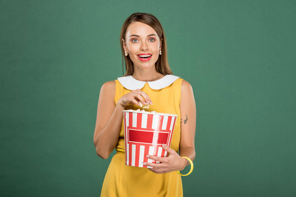 beautiful woman holding bucket of popcorn and looking at camera isolated on green - Foto, immagini