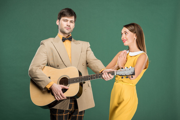 handsome man in vintage clothes playing acoustic guitar while beautiful woman doing please gesture isolated on green - Фото, изображение