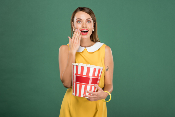 beautiful surprised woman holding bucket of popcorn and covering mouth with hand isolated on green - Foto, Imagem