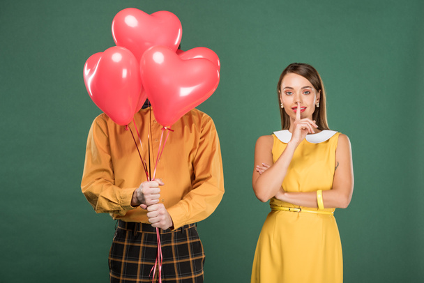 beautiful woman doing silent gesture while man covering face with heart shaped balloons isolated on green - Foto, Imagem