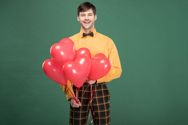 handsome smiling man in vintage clothes with heart shaped balloons isolated on green - Zdjęcie, obraz