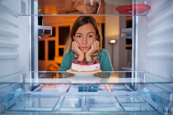 Frau, die mit dem Kopf in der Hand vor Kühlschrank steht und Käsekuchen betrachtet. Bild aus dem Inneren des Kühlschranks. - Foto, Bild