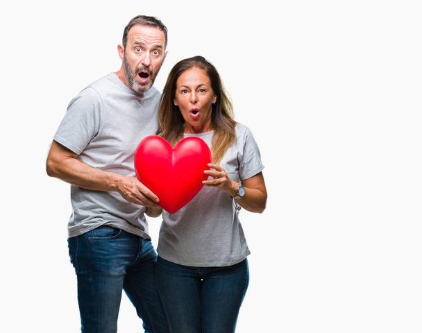 Μέση ηλικία Ισπανόφωνος casual ερωτευμένο ζευγάρι κρατώντας κόκκινη καρδιά πάνω από απομονωμένες φόντο φοβάται το σοκ με πρόσωπο έκπληξη, φοβάται και ενθουσιασμένος με φόβο έκφραση - Φωτογραφία, εικόνα
