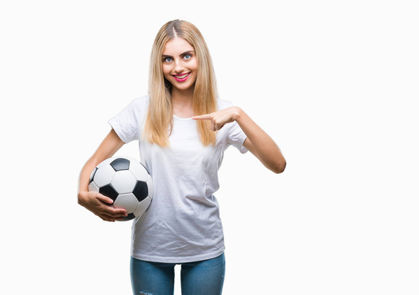 Joven hermosa mujer rubia sosteniendo pelota de fútbol sobre fondo aislado con la cara sorpresa señalando el dedo a sí mismo
 - Foto, imagen