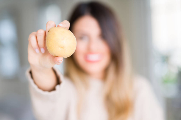 Young beautiful woman holding fresh potato at home with a happy face standing and smiling with a confident smile showing teeth - Фото, изображение