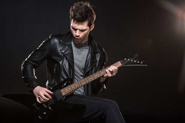 handsome rocker in leather jacket playing electric guitar on black background - Foto, Imagem