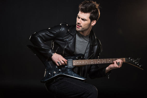 handsome rocker in leather jacket playing electric guitar and looking away isolated on black - Foto, Bild