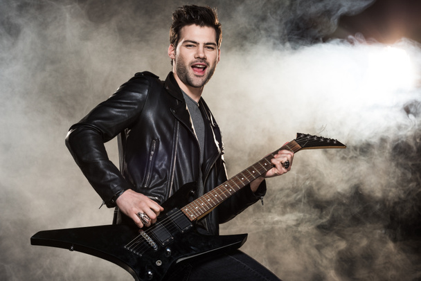 handsome rocker in leather jacket playing electric guitar on smoky background - Foto, Imagem