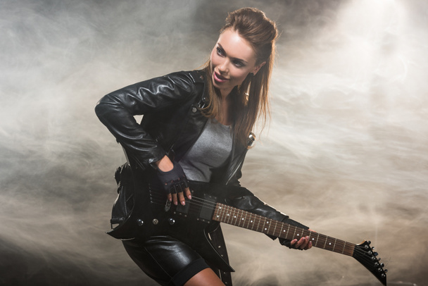 レザー ジャケットの煙のような背景にエレク トリック ギターを演奏で美しい女性 - 写真・画像