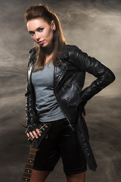 beautiful woman in leather jacket posing with electric guitar on smoky background - 写真・画像