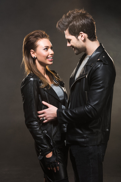 beautiful smiling couple in leather jackets looking at each other and posing on dark background - Photo, Image