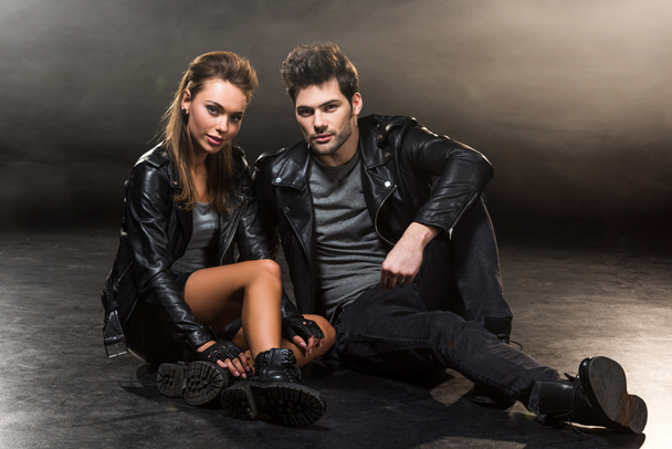 beautiful fashionable couple in leather jackets sitting and posing on dark background - Фото, изображение