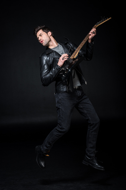 handsome rocker in leather jacket playing electric guitar isolated on black - Fotografie, Obrázek