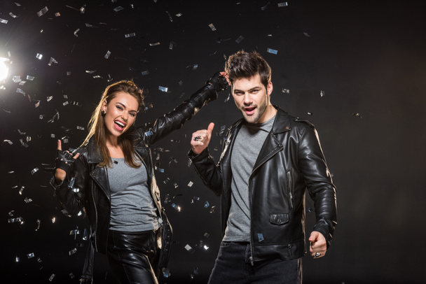 beautiful couple in leather jackets showing rock and thumb up signs while cheering with falling confetti on black background - Photo, Image