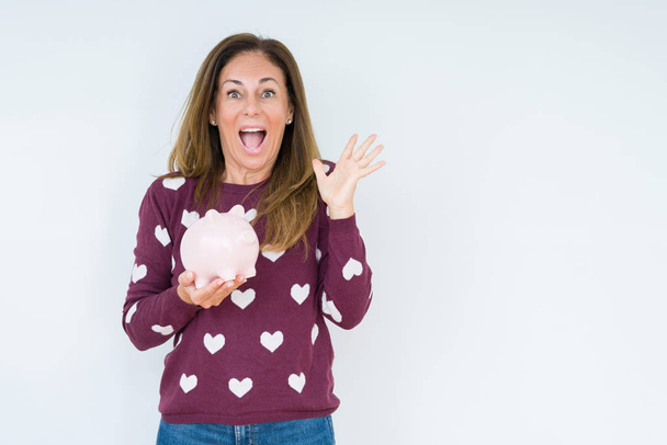 Middle age woman holding piggy bank over isolated background very happy and excited, winner expression celebrating victory screaming with big smile and raised hands - Photo, Image