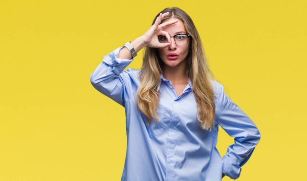 Young beautiful blonde business woman wearing glasses over isolated background doing ok gesture shocked with surprised face, eye looking through fingers. Unbelieving expression. - Photo, Image