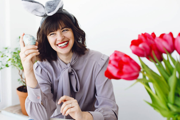 Krásná mladá žena v uši zajíček malování kraslic a usmívá se u stolu s barvy, štětce, tulipány ve váze. Šťastná dívka zdobení vajec, držení štětce, Velikonoční svátek přípravy - Fotografie, Obrázek