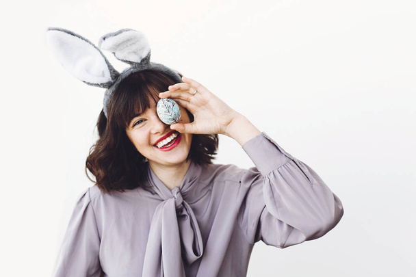 Beautiful girl in bunny ears holding easter egg near face and smiling on white background indoors, space for text. Easter hunt concept. Happy easter greetings. - Photo, Image