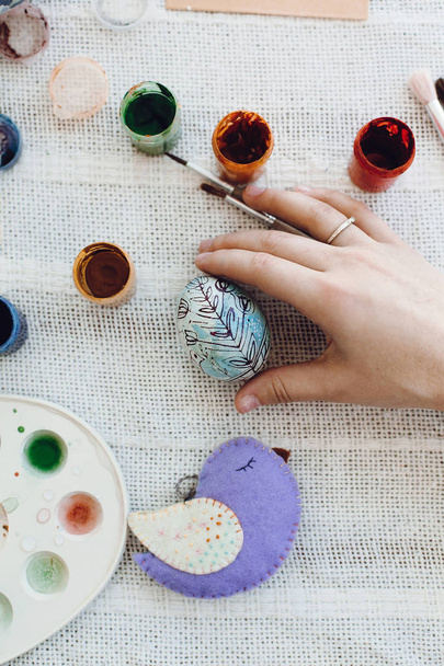 Hands holding stylish easter egg on rustic table with paint,brushes, bird toy. Flat lay , space for text. Child decorating easter egg, holiday preparations. Happy Easter concept - Photo, Image